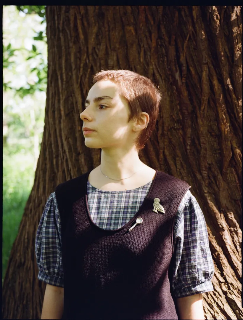 A woman with cropped hair stands in front of a tree trunk